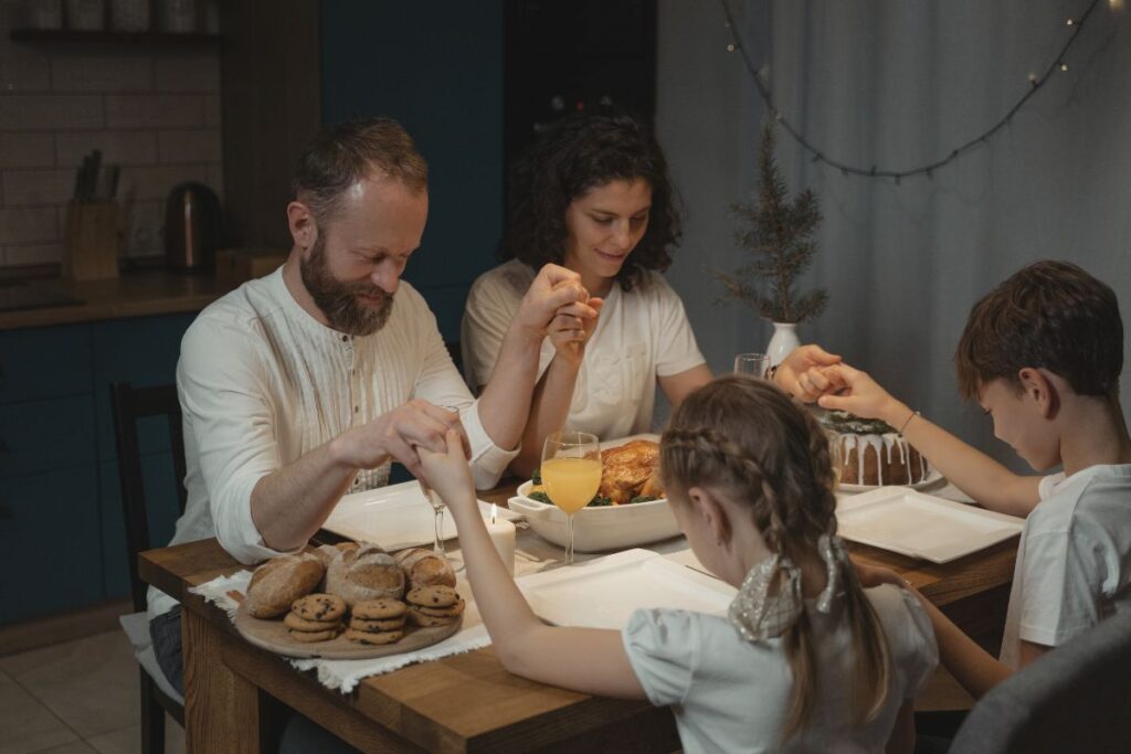 Família de quatro pessoas de mãos dadas orando ao redor de uma mesa de jantar farta.