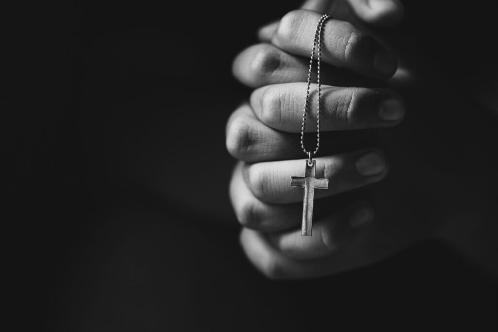 Mãos unidas em oração segurando um crucifixo de metal em fundo escuro.