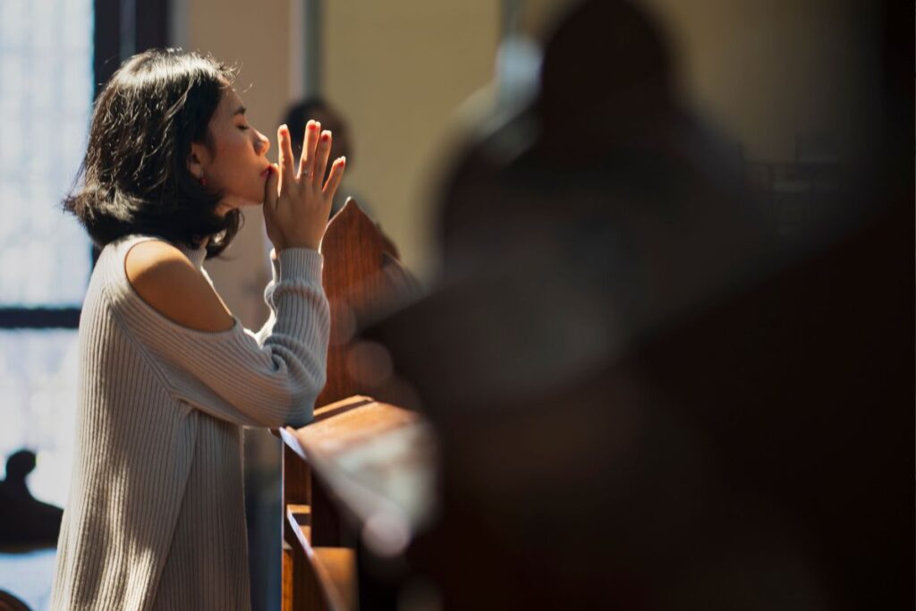 Uma mulher orando em comunidade, de joelhos em um banco de igreja com as mãos juntas em súplica.