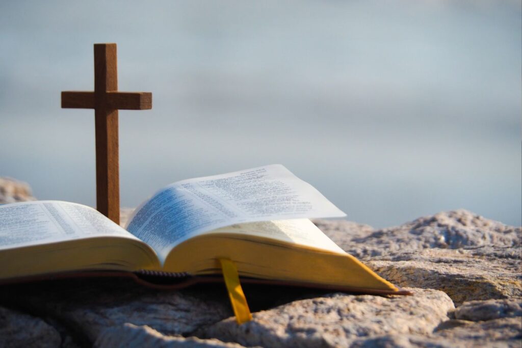 Bíblia aberta e uma cruz de madeira em uma formação rochosa, com água ou céu desfocado ao fundo, representando a perspicácia em meio às tempestades da vida.